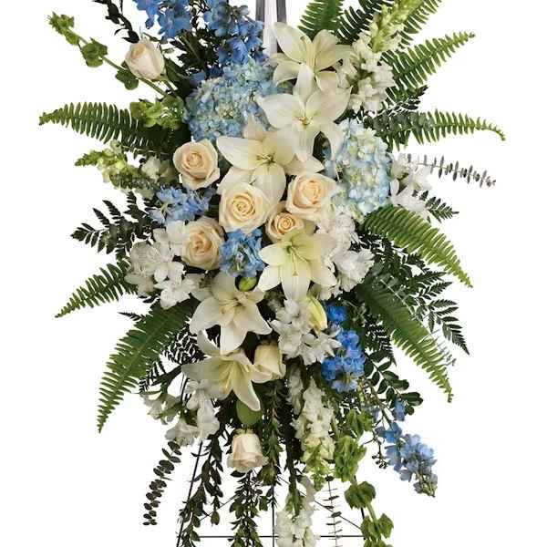 Standing funeral spray with white lilies, roses, and blue flowers on a cross easel