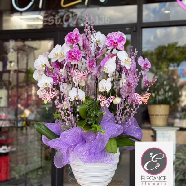 Tall arrangement of white and pink orchids in a white pot with purple mesh and butterfly accents