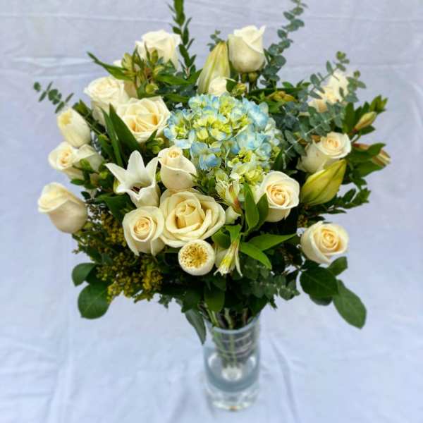 Tall glass vase of cream roses, white lilies, and blue hydrangea with mixed filler flowers.