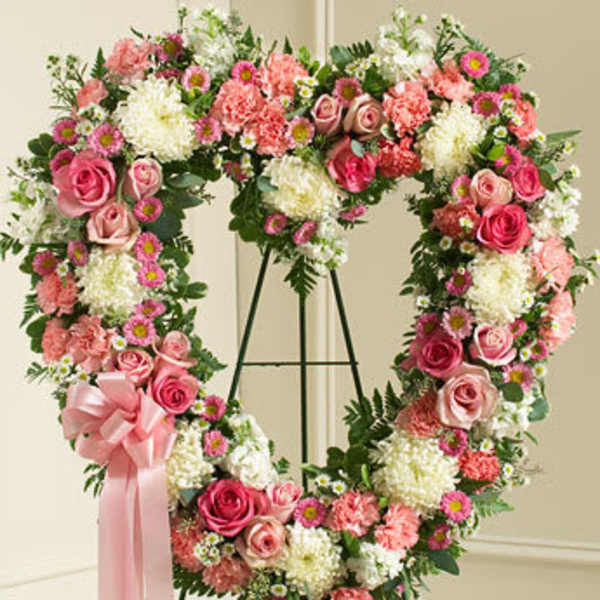 Heart-shaped standing wreath of pink roses, carnations, and white mums with a pink bow.