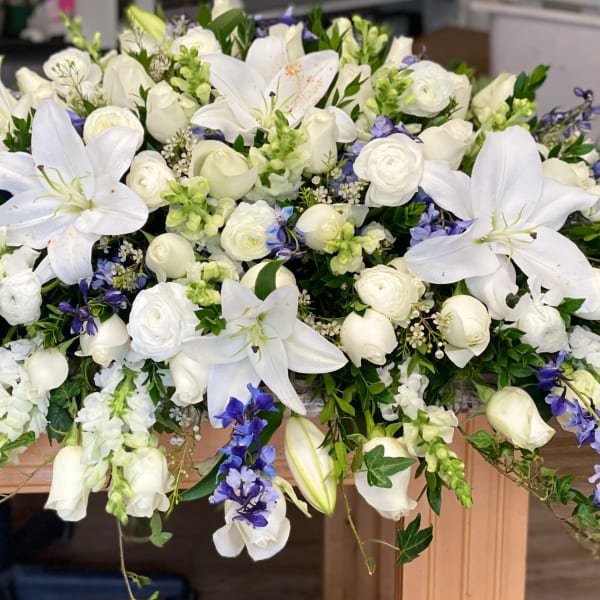 Large white floral casket spray with lilies, roses, and blue accents