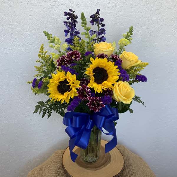 Bouquet of yellow roses, sunflowers, and purple flowers in a glass vase with a blue ribbon