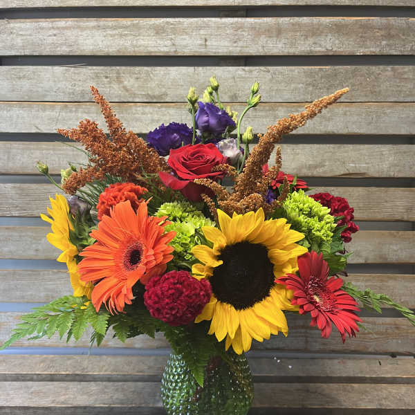 Colorful mixed bouquet with sunflowers, gerbera daisies, and roses in a green glass vase