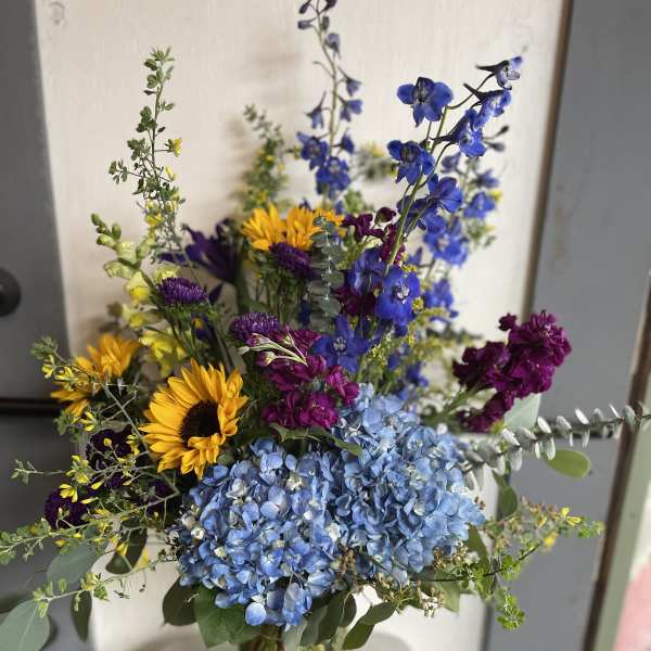 Mixed bouquet with blue hydrangeas, sunflowers, and purple blooms in a glass vase