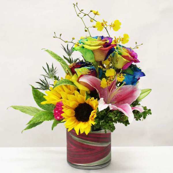 Bright mixed arrangement with rainbow roses, sunflower, pink lily and yellow orchids in a red glass vase