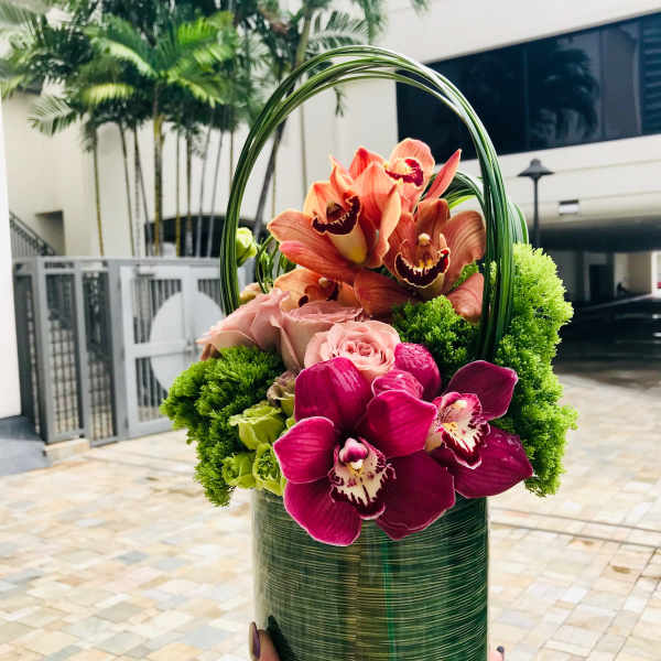 Orchid and rose arrangement in a green cylindrical container with a loop handle