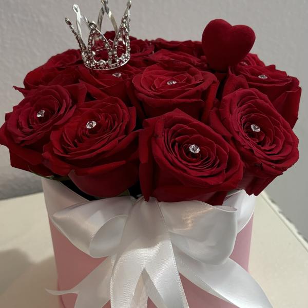 Red roses in a pink hat box with a white ribbon, crown, and heart topper