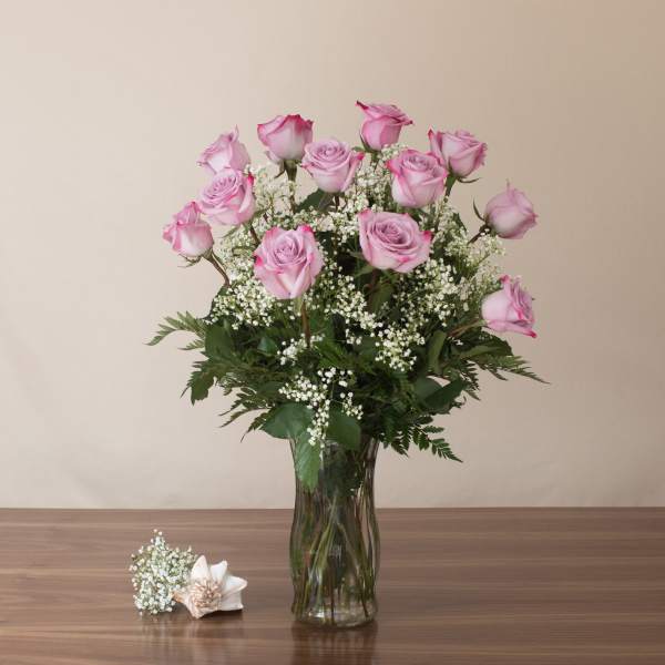 Tall glass vase of pink roses with white filler flowers and a ceramic flower on a wooden table