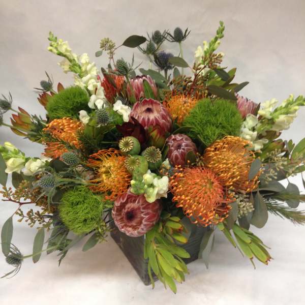 Mixed bouquet with orange pincushion protea and green accents in a dark container