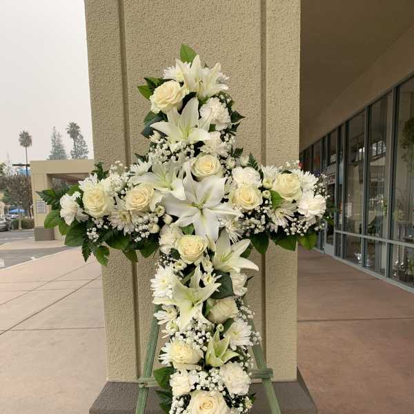 White floral cross on a stand with roses, lilies, and daisies