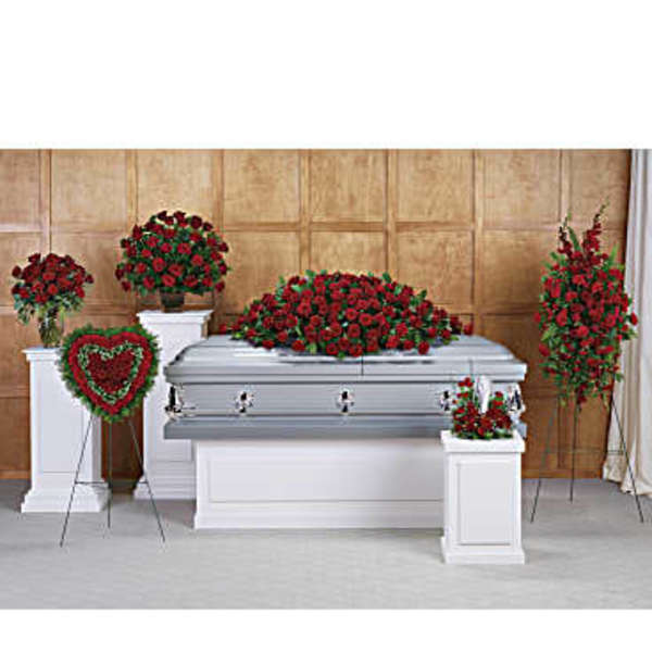 Funeral display with red rose casket spray, standing sprays, and heart wreath around a silver casket