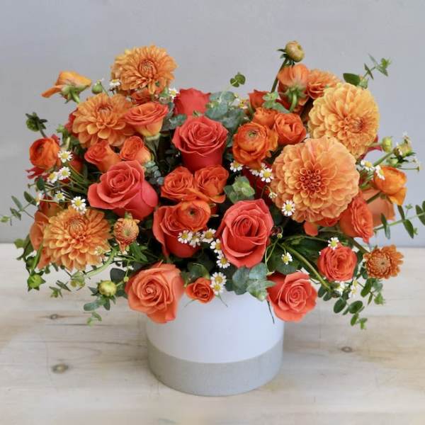 Orange roses, dahlias, and ranunculus in a low white cylinder vase with small white daisies