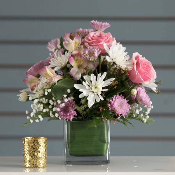 Compact arrangement of pink roses, white and lavender mums in a glass cube vase with a gold votive holder nearby