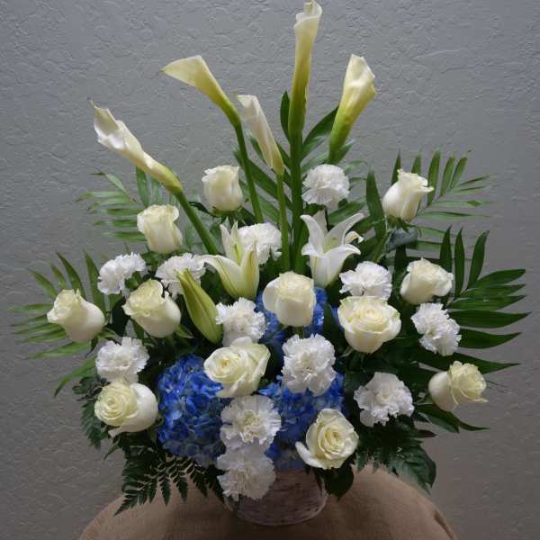 White calla lilies, roses, and carnations arranged with blue hydrangeas in a basket