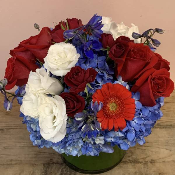 Red and white roses with blue hydrangeas and an orange gerbera daisy in a green vase