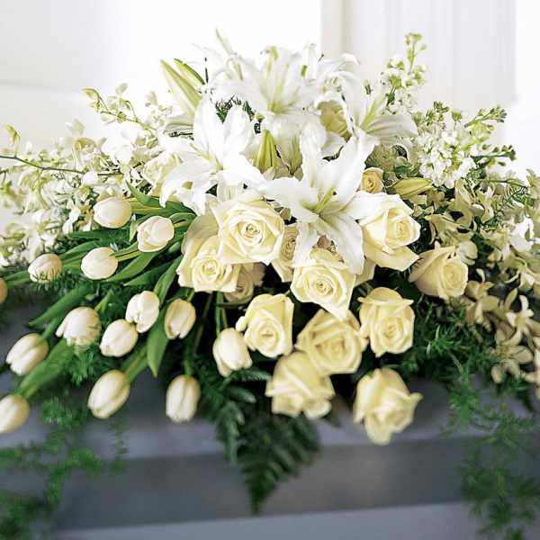Large white and cream casket spray with lilies, roses, tulips, and orchids cascading over a gray casket.
