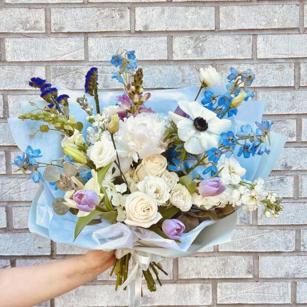 Handheld bouquet of white, blue, and lavender flowers wrapped in pale blue paper