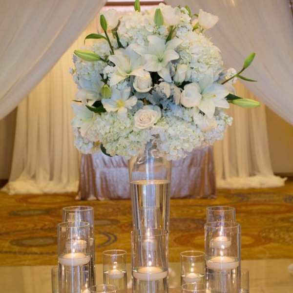 White floral centerpiece in a tall glass vase with candles around it