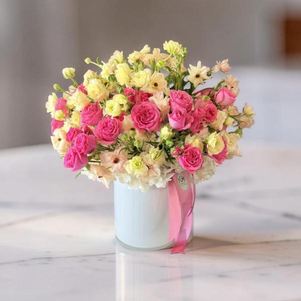Pink and cream roses arranged in a white cylindrical vase with a pink ribbon