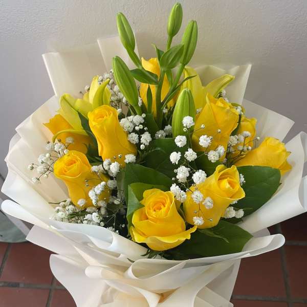 Hand-tied bouquet of yellow roses, lily buds, and white filler flowers wrapped in white paper.