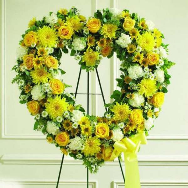 Heart-shaped standing wreath of yellow and white flowers on a metal easel with a yellow bow.