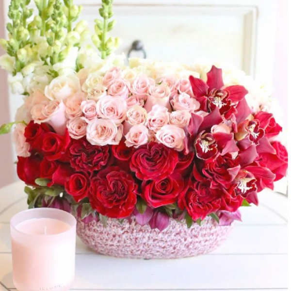 Pink and red rose arrangement in a round basket with a candle beside it.