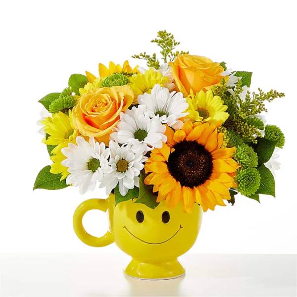 Yellow mug vase with a smiley face holding yellow, white, and orange flowers