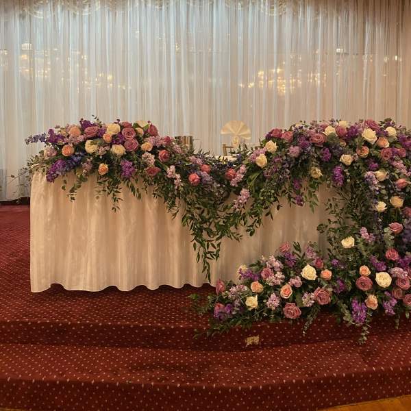 Low cascading arrangement of pink, purple, and white flowers decorating a long white table under a large chandelier