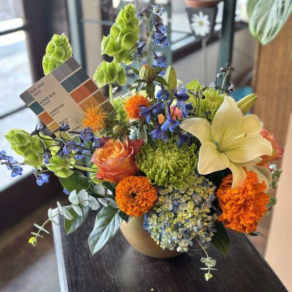 Mixed bouquet in a tan vase with orange, blue, green, and cream flowers.