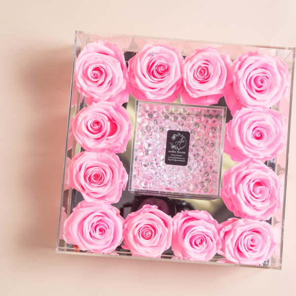 Pink roses arranged in a clear square box around a small center gift box.