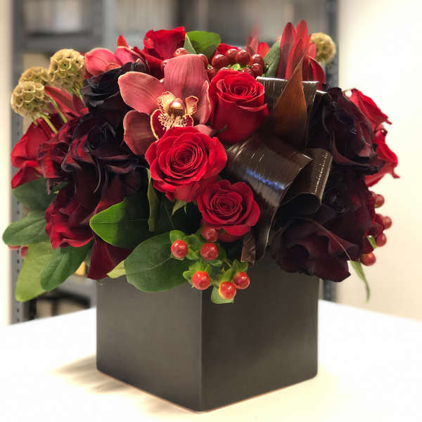 Compact arrangement of red roses and a pink orchid in a black square vase