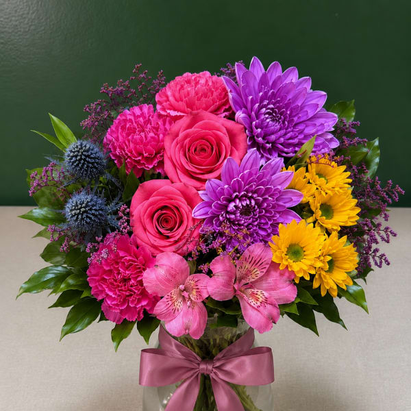 Colorful bouquet of roses, chrysanthemums, carnations, and alstroemeria in a glass vase with a pink ribbon