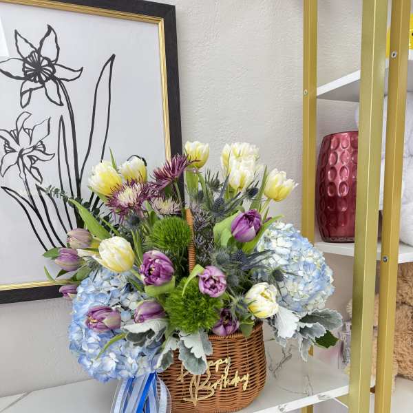 Basket arrangement of purple, white, and blue flowers with a birthday ribbon