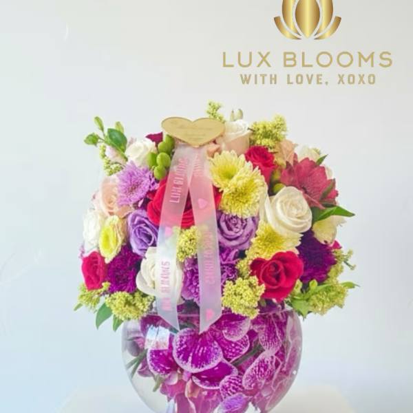 Colorful mixed flower arrangement in a round glass vase with a pink ribbon