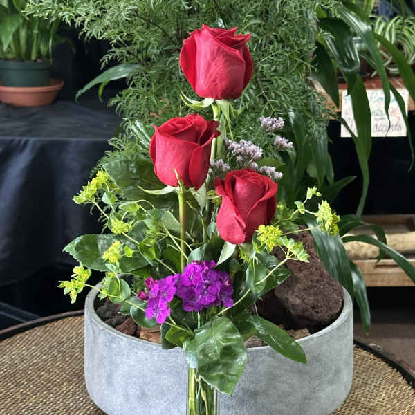 Three red roses in a clear glass vase with purple and yellow accents