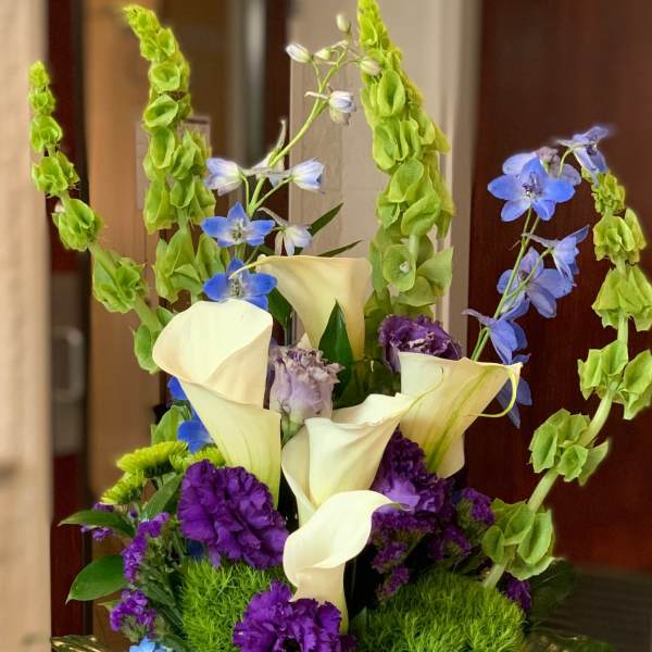 Tall bouquet of white calla lilies, blue flowers, and purple blooms in a glass vase