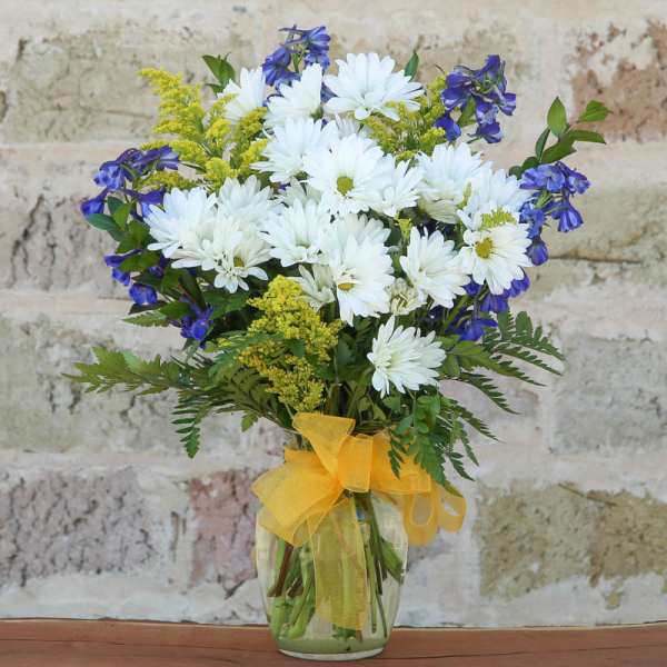 White daisies and blue flowers in a glass vase with a yellow ribbon