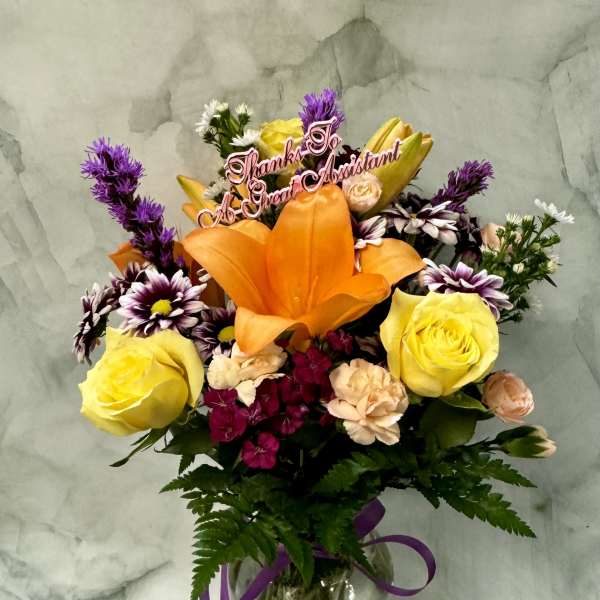 Mixed bouquet with orange lily, yellow roses, and daisies in a glass vase with a thank-you pick.