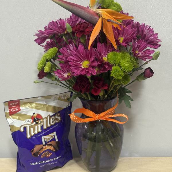 Purple and green flower arrangement in a glass vase with a chocolate candy bag