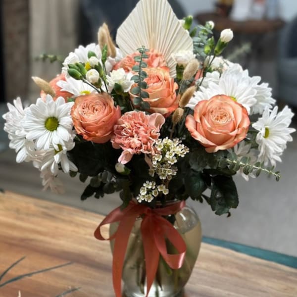 Peach roses and white daisies arranged in a glass vase with a red ribbon