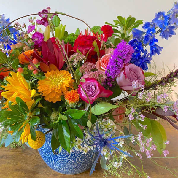 Colorful mixed flower arrangement with roses, delphinium, and daisies in a blue patterned ceramic pot.