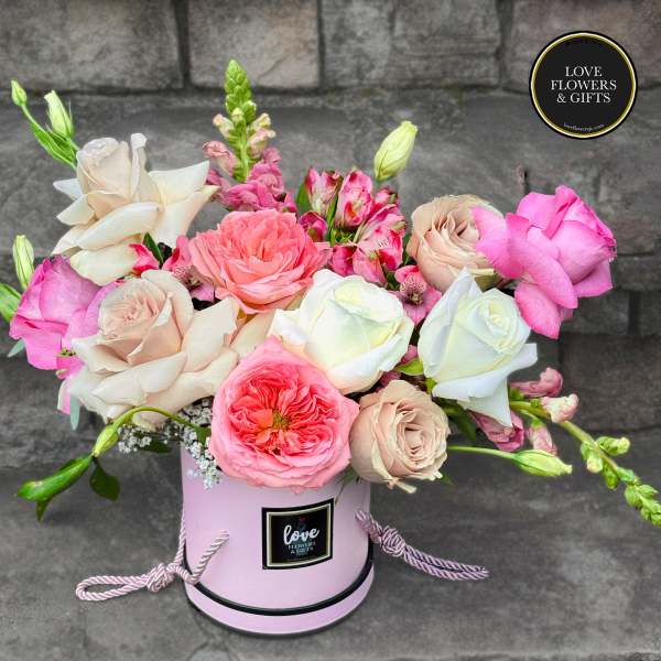 Pink and cream roses with mixed blooms arranged in a round pink hatbox-style container.