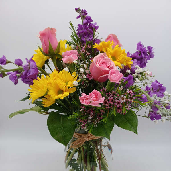 Mixed bouquet of pink roses, yellow daisies, and purple flowers in a glass vase