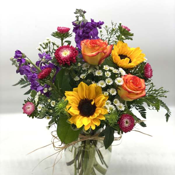 Mixed bouquet with sunflowers, orange roses, purple flowers, and white daisies in a glass vase