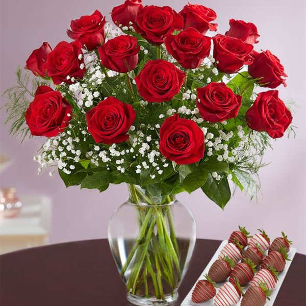 Red roses in a glass vase beside a tray of chocolate-covered strawberries