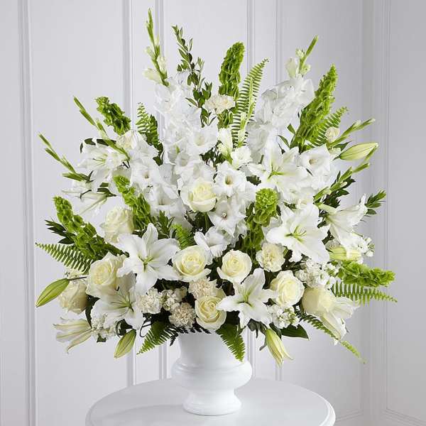 Tall white arrangement of lilies, roses, and carnations with green accents in a white urn vase