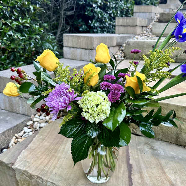 Mixed bouquet with yellow roses, purple blooms, and blue irises in a glass vase