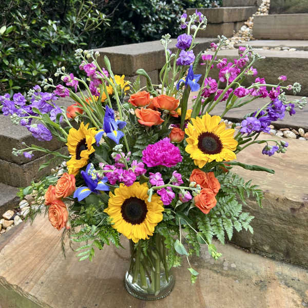 Colorful mixed bouquet in a clear glass vase with sunflowers, roses, and purple blooms