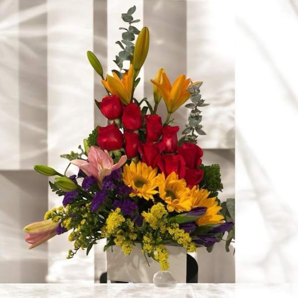 Mixed bouquet with red roses, yellow lilies, and sunflowers in a white container