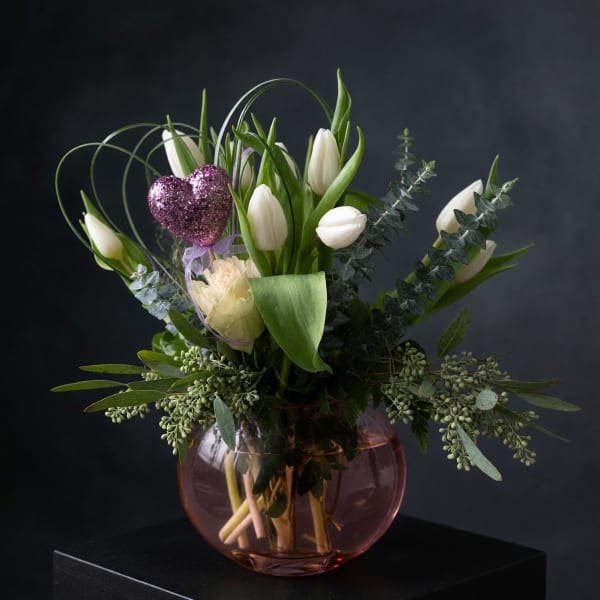 White tulips arranged in a pink glass vase with a glittery heart pick